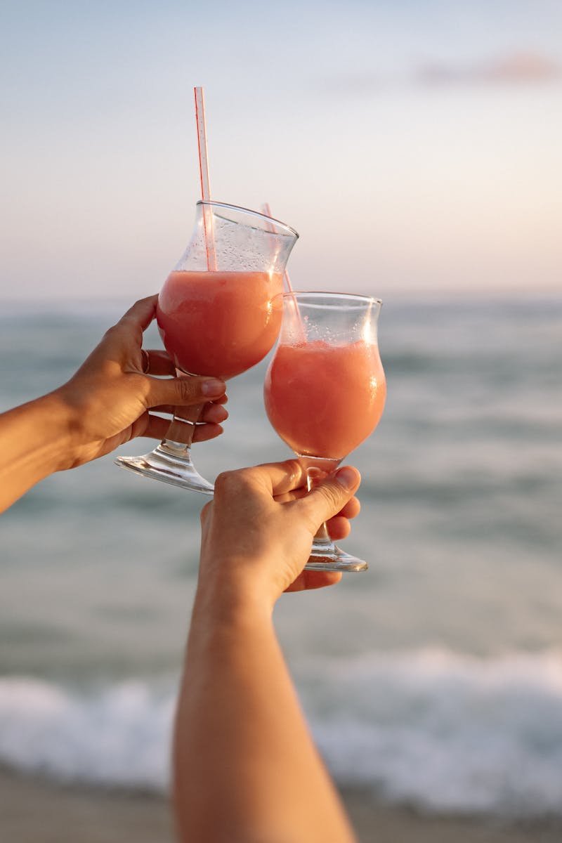 Two people celebrate with tropical cocktails by the ocean at sunset, embodying summer relaxation.